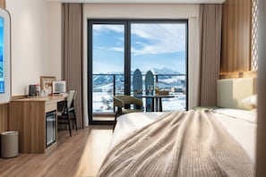 Habitación de hotel con cama, escritorio, silla, una mesita y vistas a montañas a través de amplias ventanas.