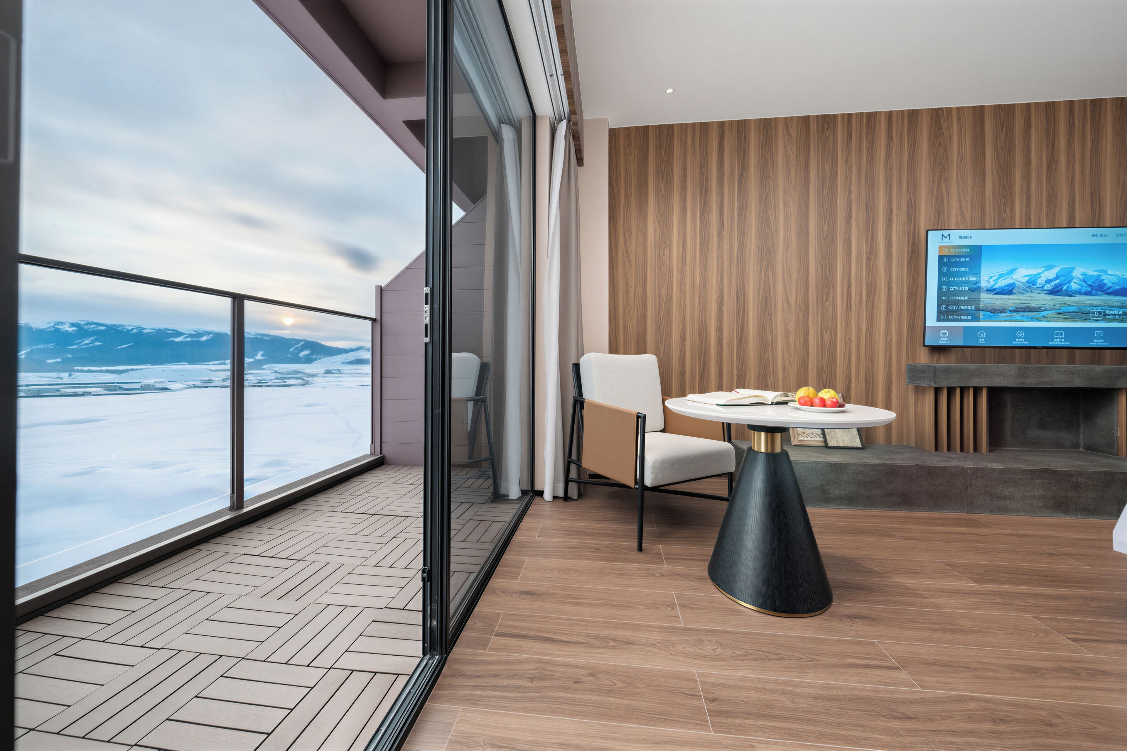 Una sala de estar moderna con un televisor grande, una mesa redonda con frutas y vistas a montañas a través de amplios ventanales.