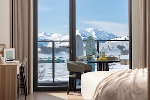 Ein Hotelzimmer mit großem Fenster, einem Bett, einem Schreibtisch mit Stühlen und einem Tisch mit Surfbrett und Flaschen.