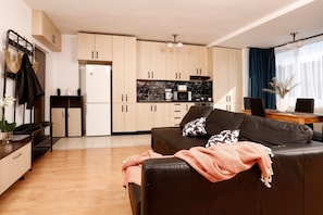 Ein modernes Wohnzimmer mit einer schwarzen Couch, einem Esstisch mit Stühlen, einer Küchenzeile mit Schränken und Geräten sowie einem großen Fenster mit Vorhängen.