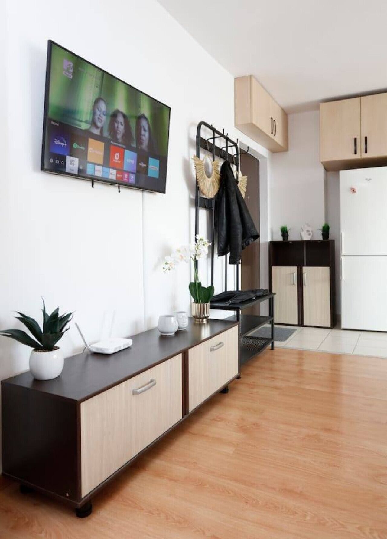 A modern living room with a flat-screen TV mounted on the wall, a wooden cabinet with drawers, a small table with a potted plant, and a kitchen area with cabinets and a refrigerator.