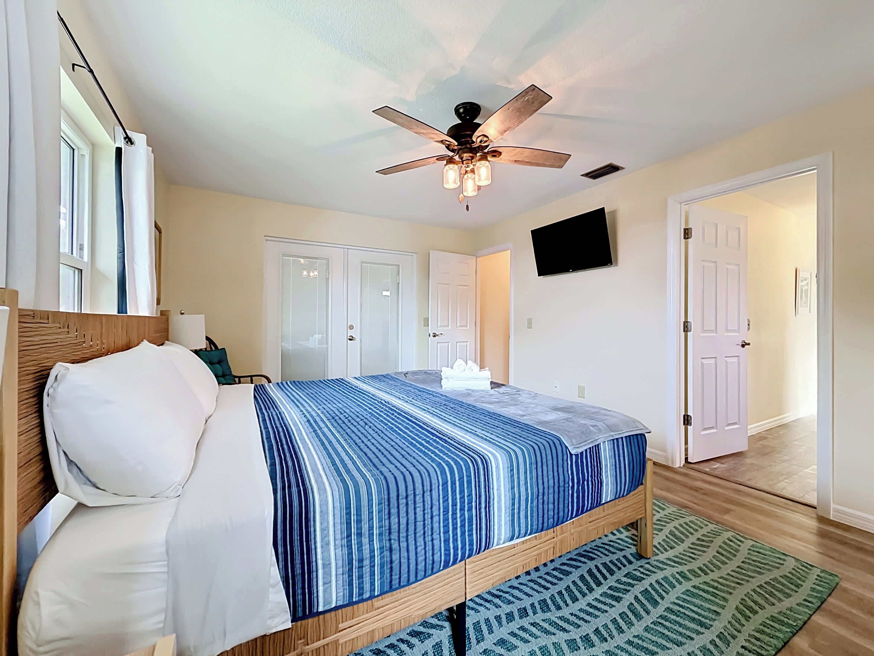 A bedroom with a bed, a ceiling fan, a TV, and a door leading to another room.