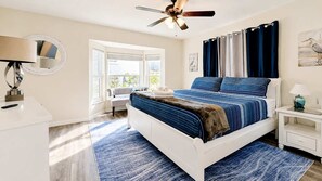 Ein Schlafzimmer mit einem Bett, einem Schreibtisch, einem Stuhl, einem Fenster und einem Deckenventilator.