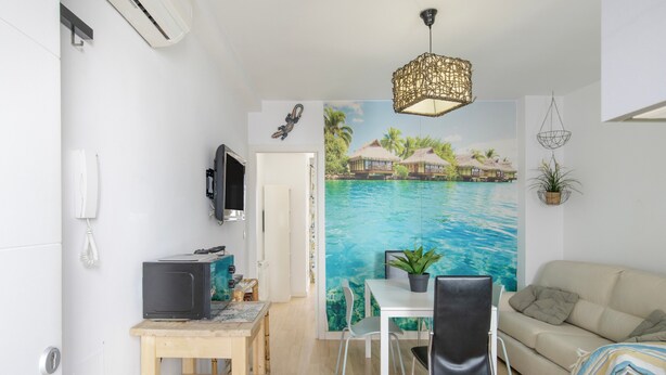 Espai vital reduït amb zona de menjador, un sofà, un televisor i un mural d'una platja tropical.