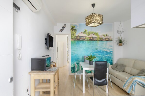 A compact living space with a dining area, a sofa, a TV, and a mural of a tropical beach. - Central Polynesia Apartment in Ronda (Ronda)
