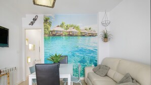 A room with a dining table, chairs, a sofa, a TV, a wall mural of a tropical beach, and a small plant. - Central Polynesia Apartment in Ronda (Ronda)