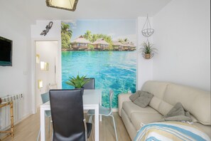 Una habitación con una mesa de comedor, sillas, un sofá, un televisor, un mural en la pared de una playa tropical y una planta pequeña.