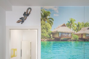 Una habitación con una puerta, un adorno en la pared y un gran ventanal con vistas a bungalows sobre el agua y palmeras.