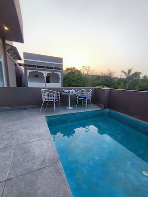 Una piscina en la azotea con dos sillas blancas y una mesa, rodeada por un muro bajo y un edificio de arquitectura tradicional.