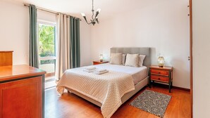 Quarto com cama, mesas de cabeceira, escrivaninha, lustre e vista para o verde lá fora.