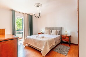 Une chambre à coucher avec un lit, des tables de chevet, un bureau, un lustre et une vue sur la verdure à l’extérieur.