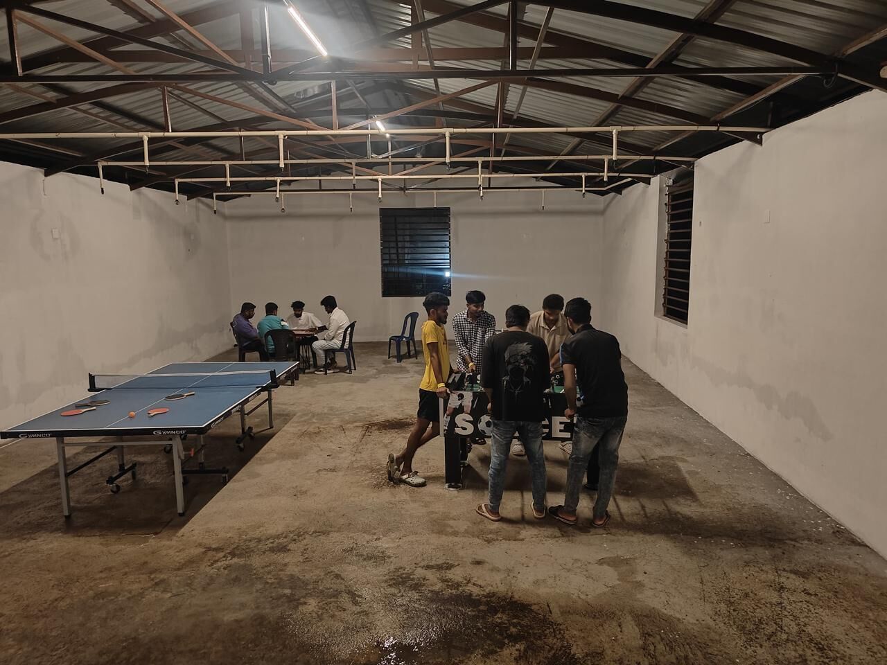 A room with a ping pong table, chairs, and a group of people.