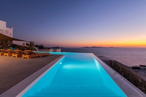 Zona de piscina con sillones de descanso y vista al mar al atardecer.