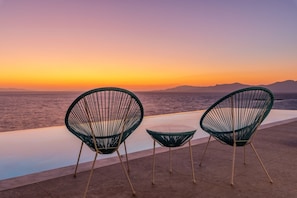 Twee groene wireframe stoelen en een kleine tafel op een balkon met uitzicht op een wateroppervlak tijdens zonsondergang.