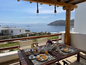 Mesa de madeira posta para o pequeno-almoço, com vista para a costa, edifícios e o mar.