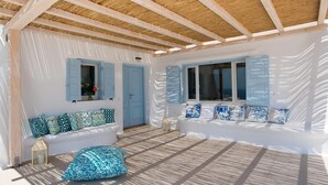 Een kamer met een houten plafond, witte muren en blauwe luiken. Er staat een witte bank met gekleurde kussens en een passende voetenbankje.