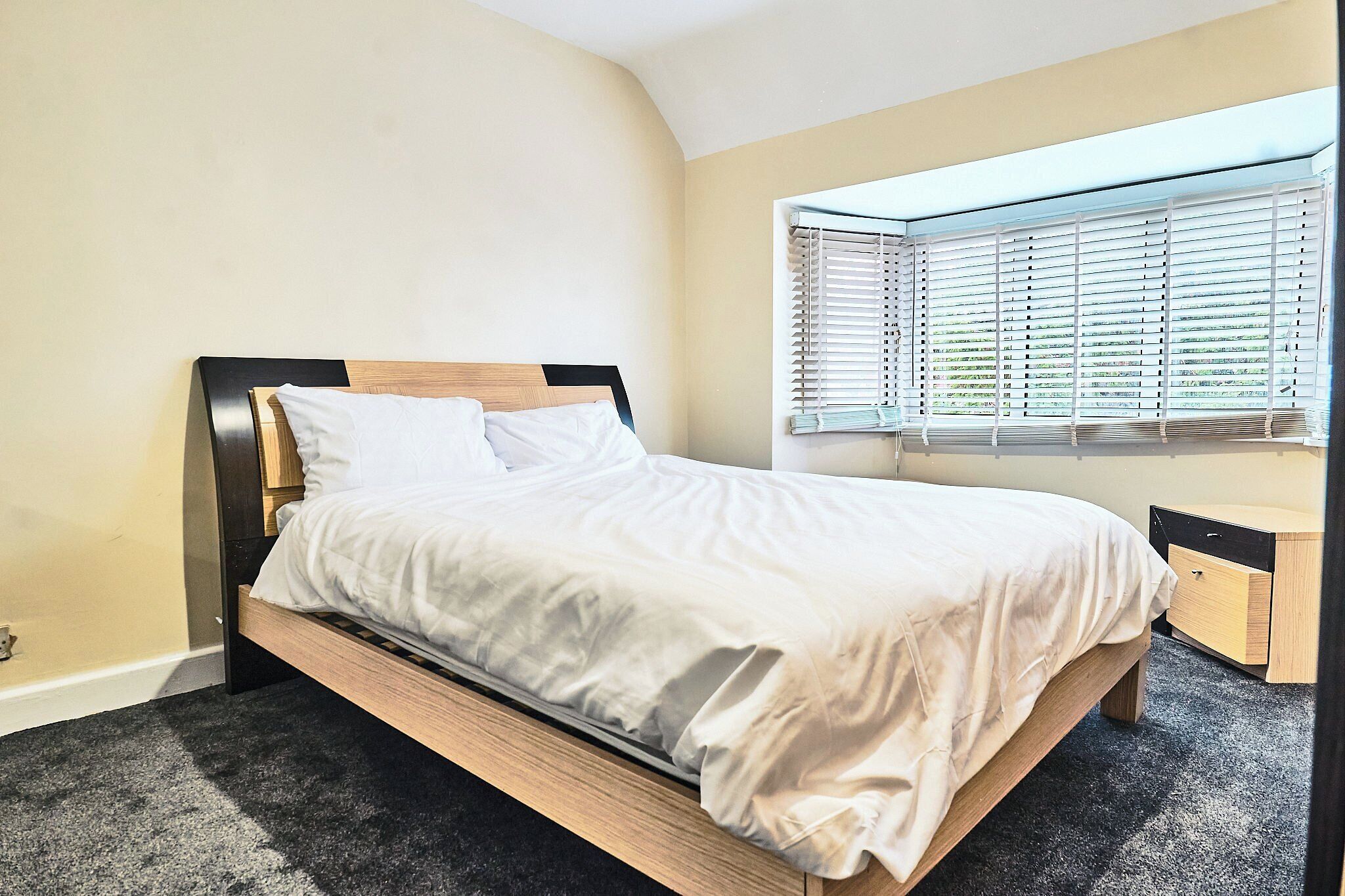 A bed with a wooden frame and white bedding, a bedside table, a window with blinds, and a carpeted floor.