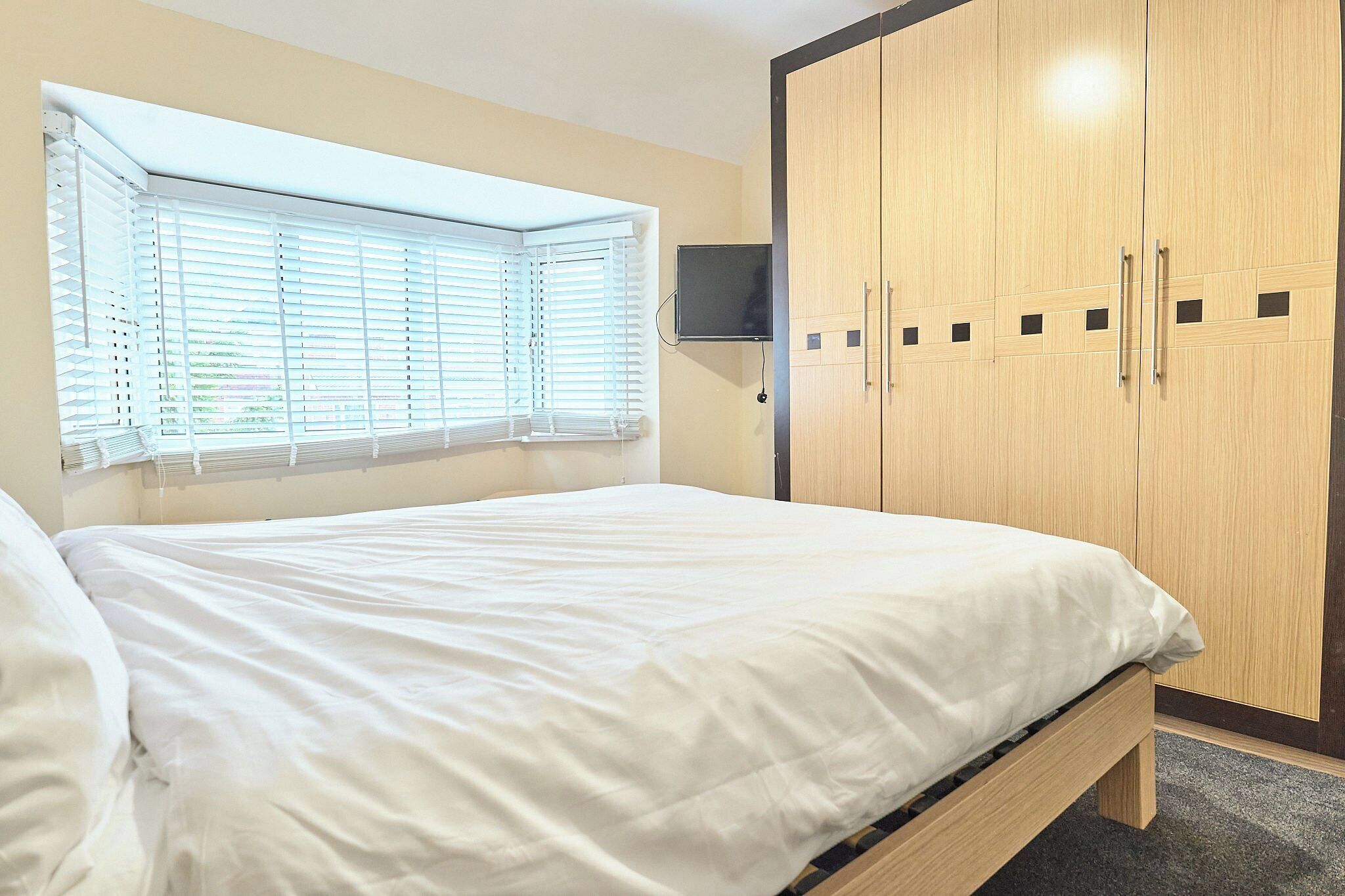A bedroom with a bed, a window with blinds, a wardrobe, and a flat-screen TV mounted on the wall.
