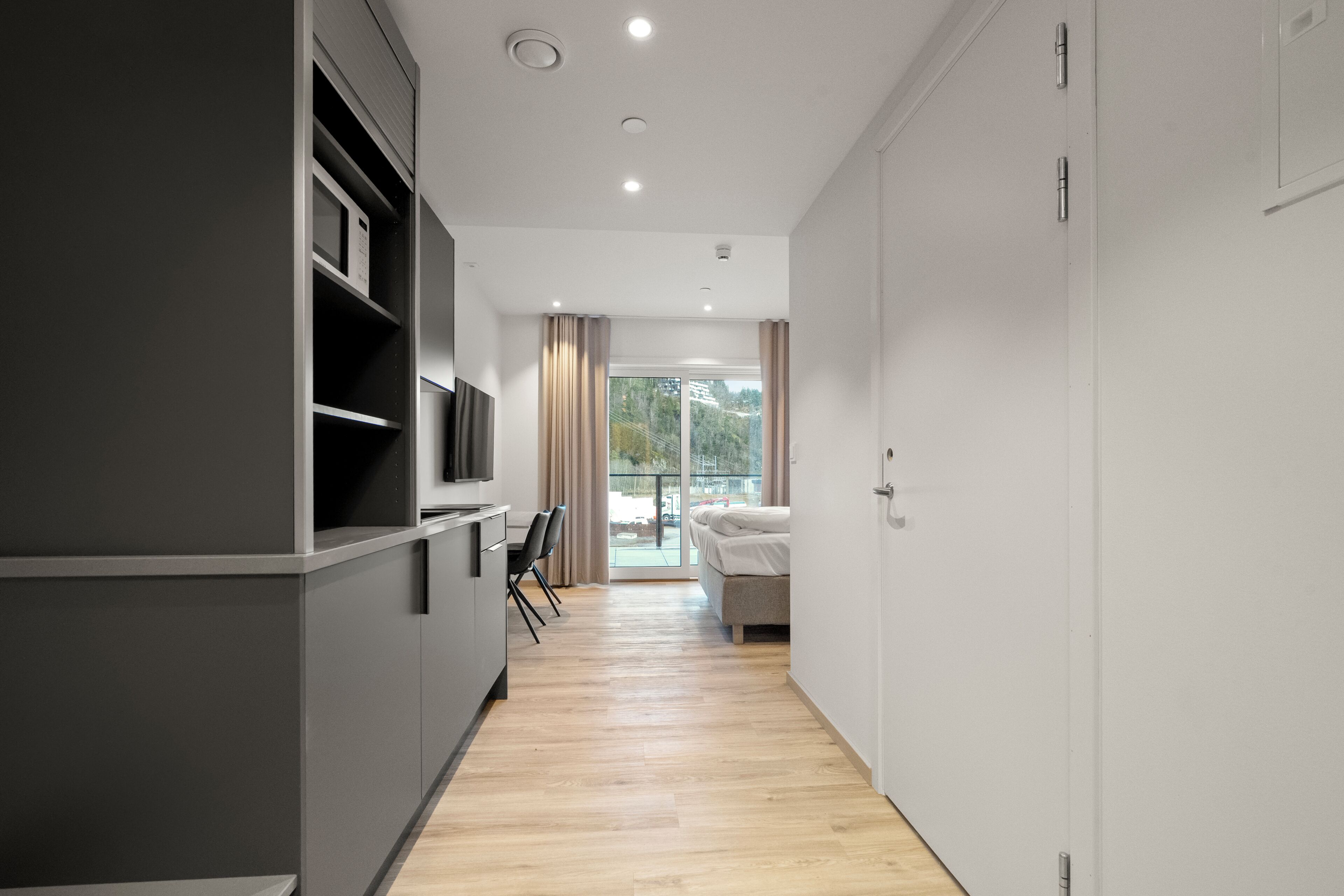 A modern hotel room with a kitchenette, a bed, a seating area, and a large window.