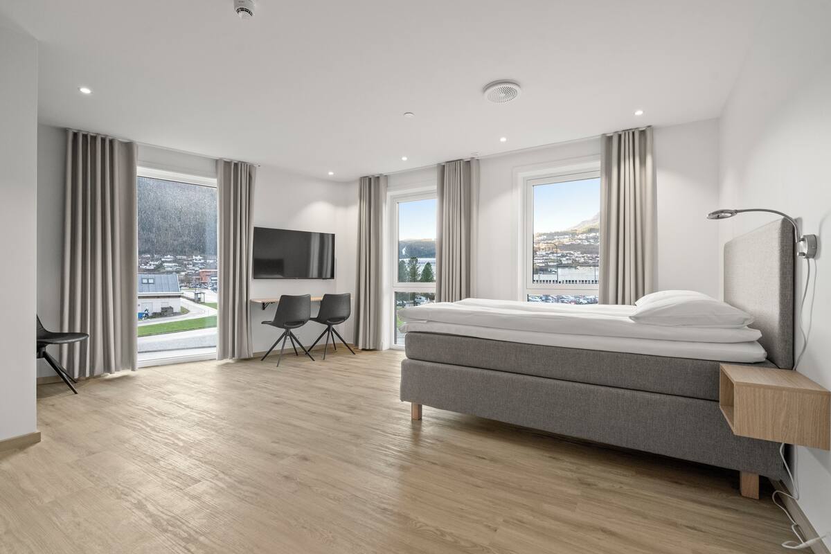 A modern hotel room with a large bed, a TV, a desk with two chairs, and a view of a cityscape.