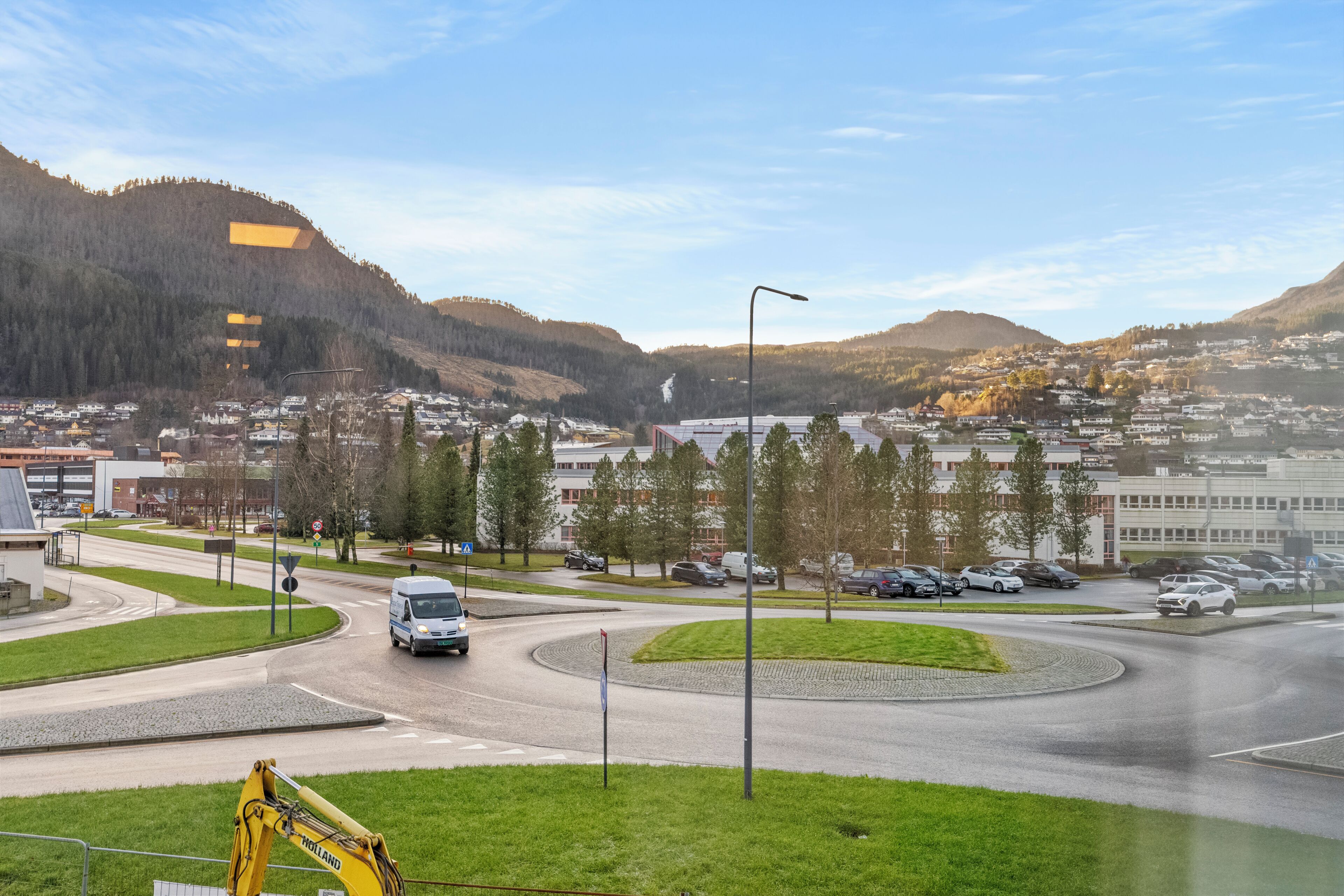 Una glorieta con una excavadora amarilla, una camioneta blanca y un área residencial con edificios y árboles al fondo.