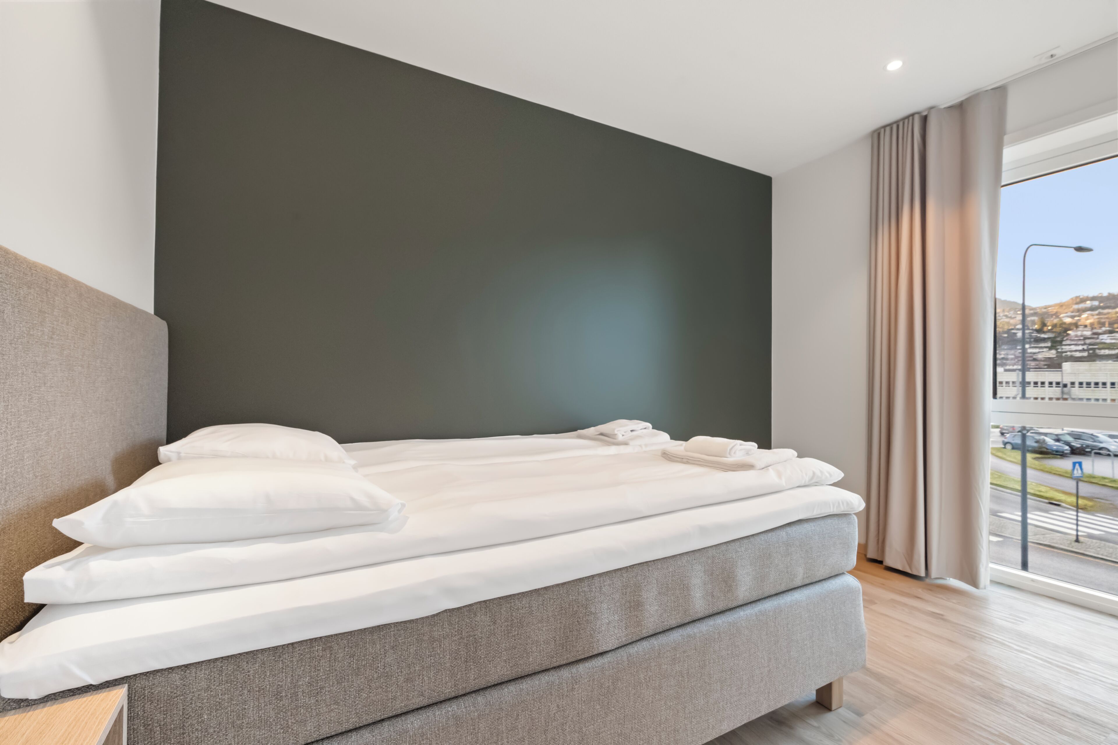 Una habitación de hotel moderna con una cama grande, una pared decorativa oscura y vistas a un paisaje urbano a través de amplios ventanales.
