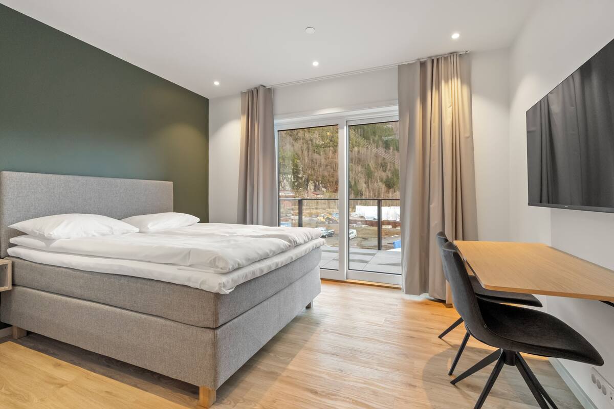 A modern bedroom with a bed, a desk, and a view through a sliding glass door.