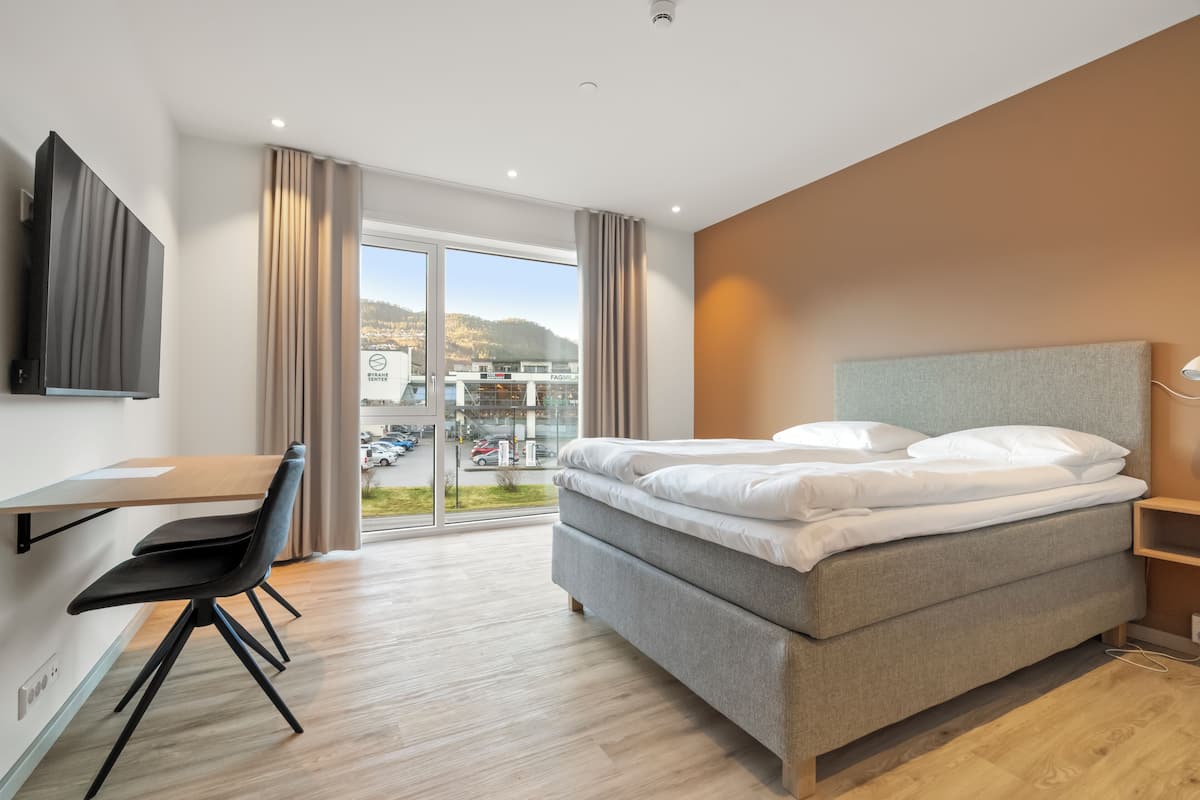 A modern hotel room with a bed, desk, chair, and a large window offering a view of the city.