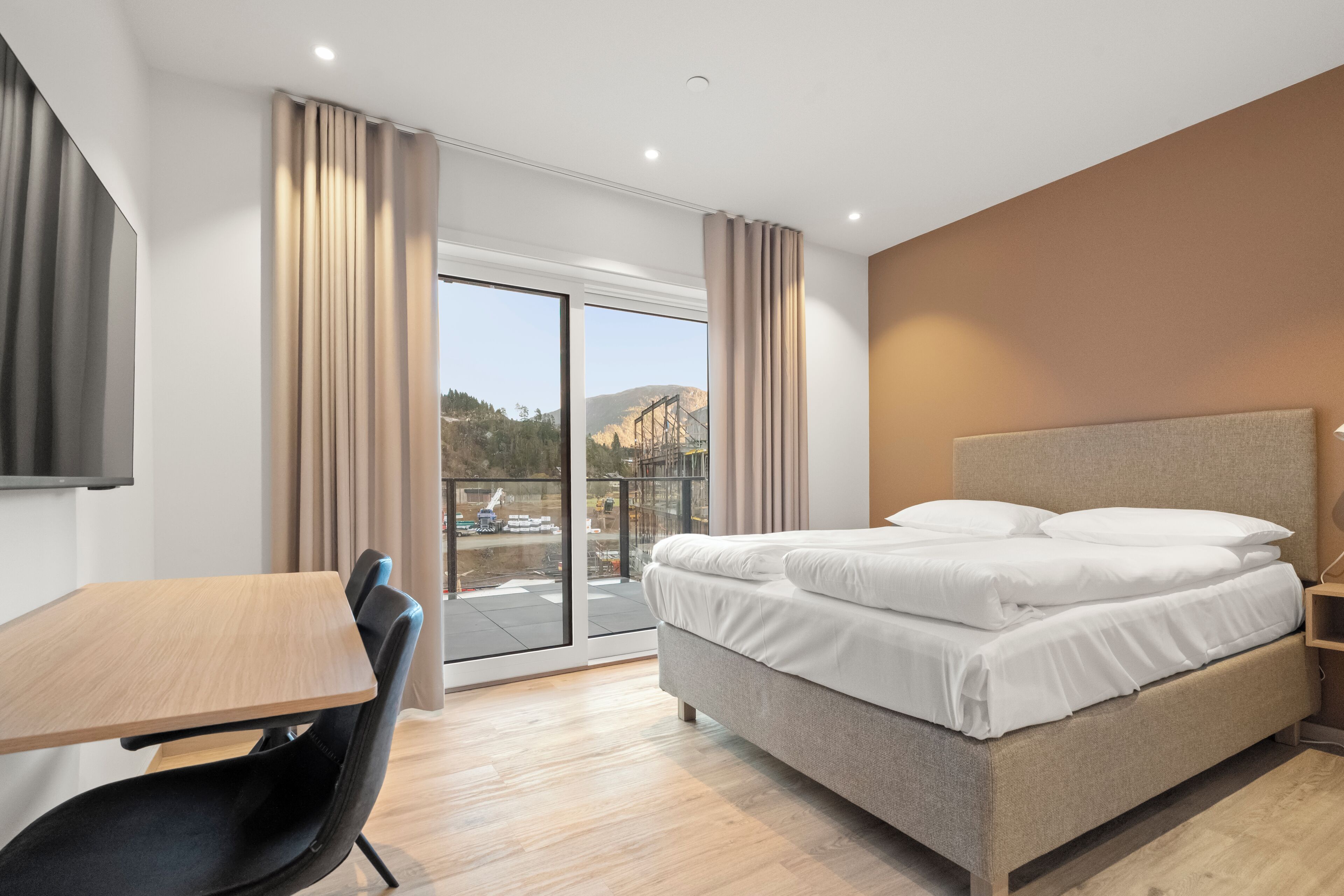A modern hotel room with a bed, a desk, and a view of a mountainous landscape.