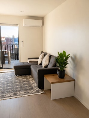 A modern living room with a grey sofa, a small wooden side table, a potted plant, and a patterned rug. - Stylish Central Christchurch Apartment | Prime Location & Comfort (Christchurch)