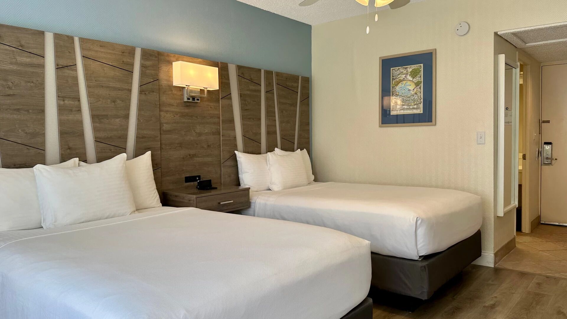 Habitación de hotel con dos camas, una mesita de noche, una lámpara de pared y una puerta que da a otra habitación.