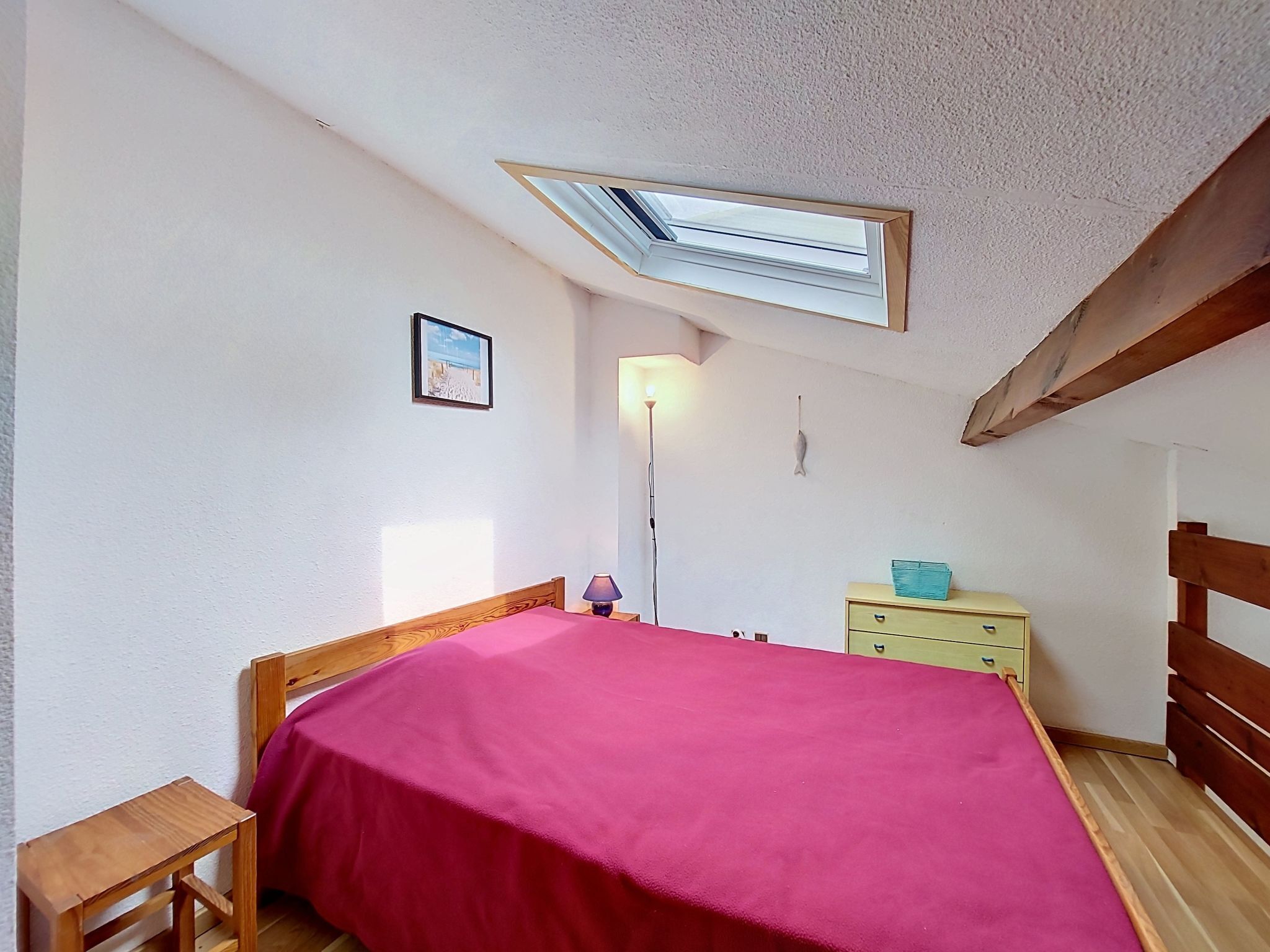 A bedroom with a skylight, wooden bed frame, a bedside table, a lamp, and a dresser.