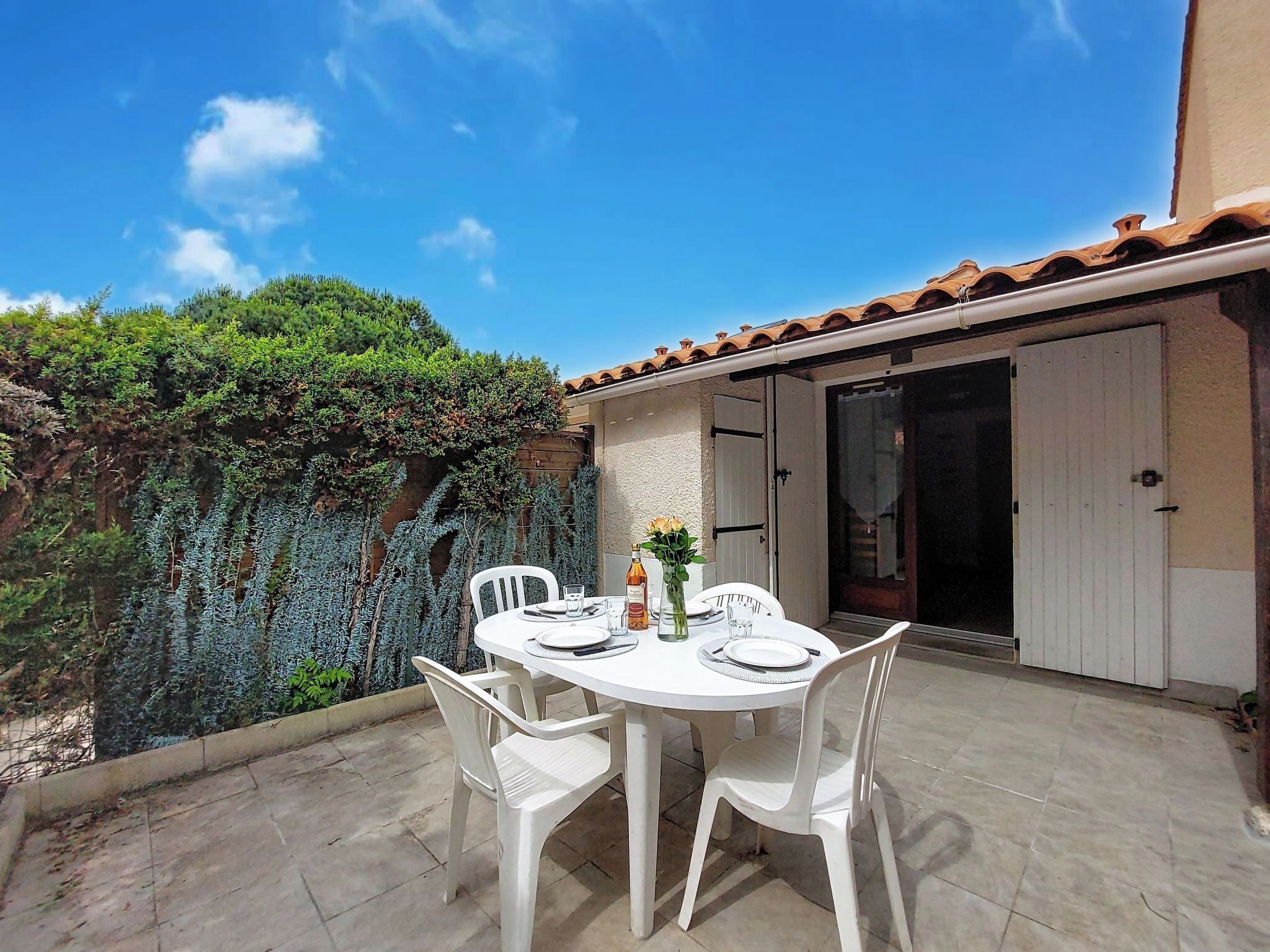 Un patio con una mesa preparada para dos personas, rodeado de sillas blancas, un suelo de baldosas y una puerta que da a una zona cubierta.