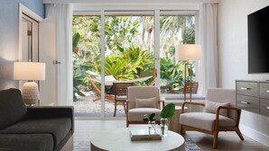 En moderne stue med sofa, to lænestole, sofabord og fjernsyn. Der er en stor skydedør, der fører ud til en have med tropiske planter og en hængekøje.