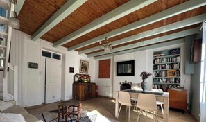 Un vaste salon doté d’un haut plafond, de poutres apparentes, d’un coin repas avec des chaises, d’une bibliothèque et d’une cheminée.