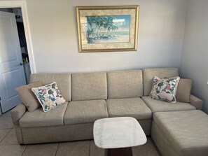 Ein beiges Ecksofa mit floralen Kissen, ein weißer Hocker und ein gerahmtes Strandbild an der Wand.