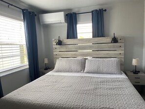 Ein Schlafzimmer mit Holzkopfstütze, einem Bett mit weißer Bettwäsche, zwei Nachttischen mit Lampen und einer Klimaanlage oberhalb der Kopfstütze.