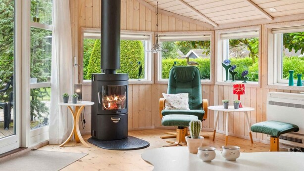 Ein gemütlicher Raum mit Kaminofen, einem bequemen Sessel, einem kleinen Tisch mit Vase und großen Fenstern mit Blick ins Grüne.
