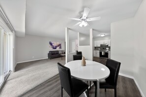 Una sala de estar moderna con un ventilador de techo blanco, una mesa de comedor con cuatro sillas, un sofá y una cocina al fondo.