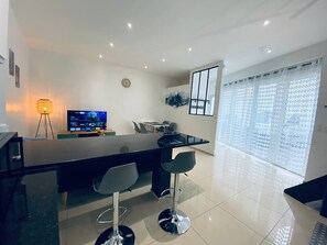 Uma cozinha moderna com bancada preta, dois bancos altos, uma mesa de jantar, uma TV e uma janela com cortinas estampadas.