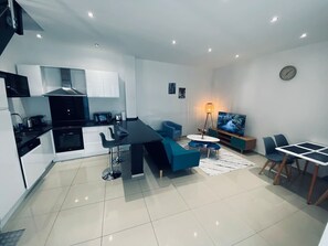 Cozinha moderna com armários brancos, bancada preta e forno embutido. Há um sofá azul e uma pequena mesa redonda com cadeiras. Uma televisão de tela plana está fixada na parede.