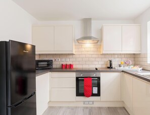 A modern kitchen with a black refrigerator, a microwave, a stove, and a red oven mitt hanging on the oven handle. - Birmingham Estate | Ultimate Summer Getaway (Winson Green)