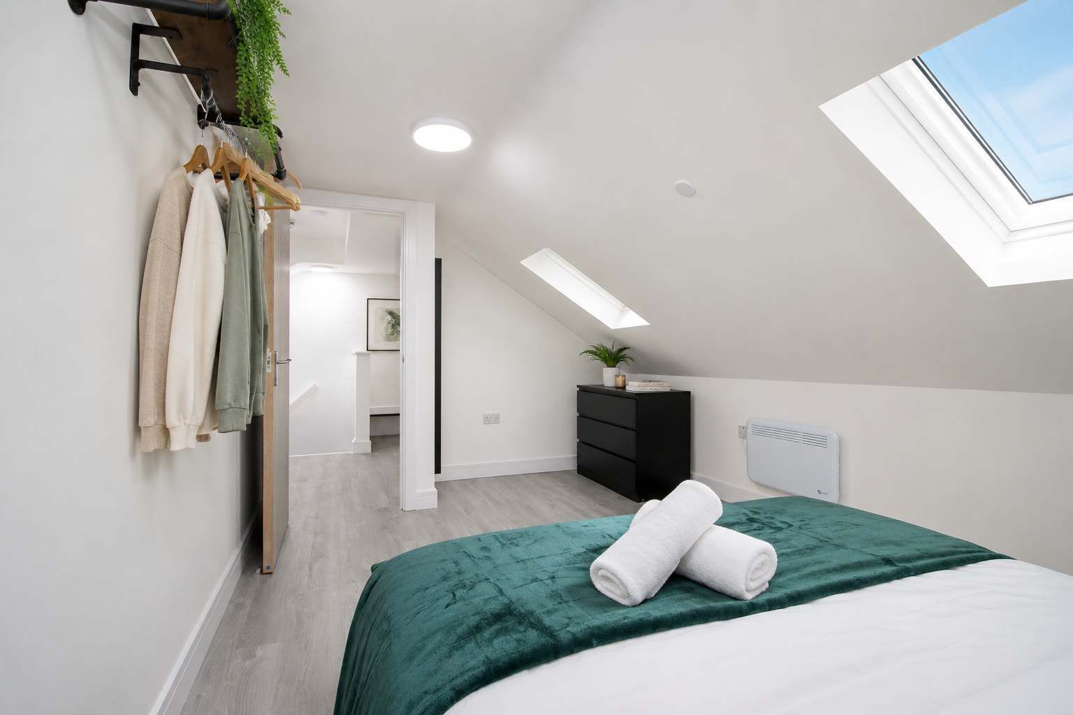 Une chambre avec une lucarne, un lit recouvert d’une couverture verte, une commode noire et une plante posée sur la commode.