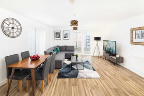 Uma sala de estar moderna com sofá, mesa de jantar, cadeiras, mesa de centro, relógio, obras de arte, televisão e abajur.
