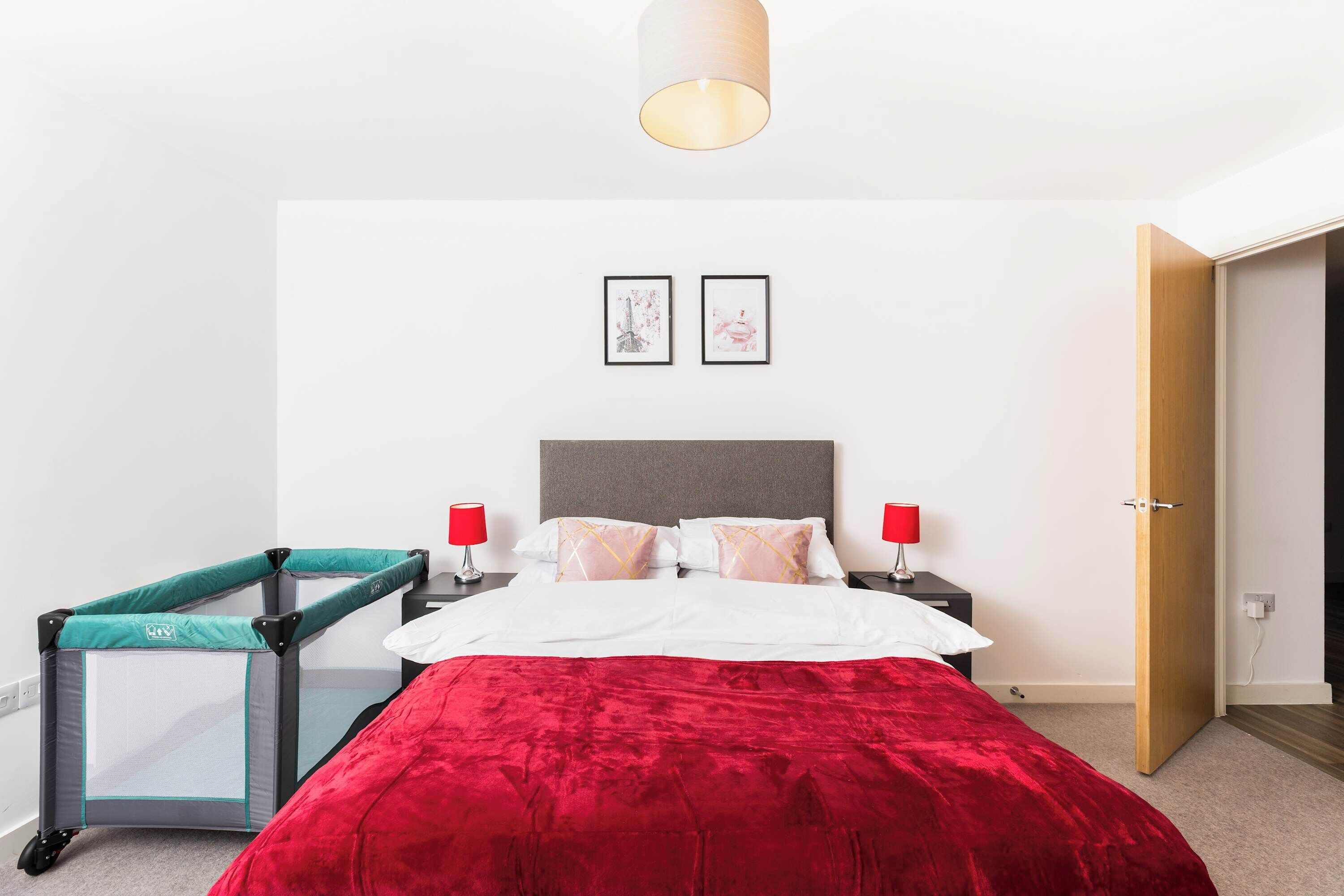 A bedroom with a bed, a red blanket, a small playpen, two framed pictures on the wall, and a door.