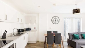 Eine moderne Küche mit weißen Schränken, ein Essbereich mit Tisch und Stühlen, ein graues Sofa mit grünen Kissen und eine große Wanduhr.
