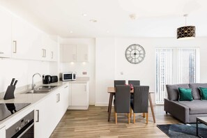A modern kitchen with white cabinets, a dining area with a table and chairs, a gray sofa with green cushions, and a large wall clock. - The Lincoln Tribe | 3 Bed Group Getaway | Parking (West Midlands)