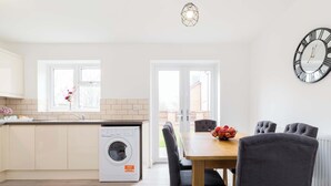A modern kitchen with a built-in washing machine, a dining area with a table and chairs, and a large wall clock. - Birmingham Estate | Made For Group Memories (Birmingham)