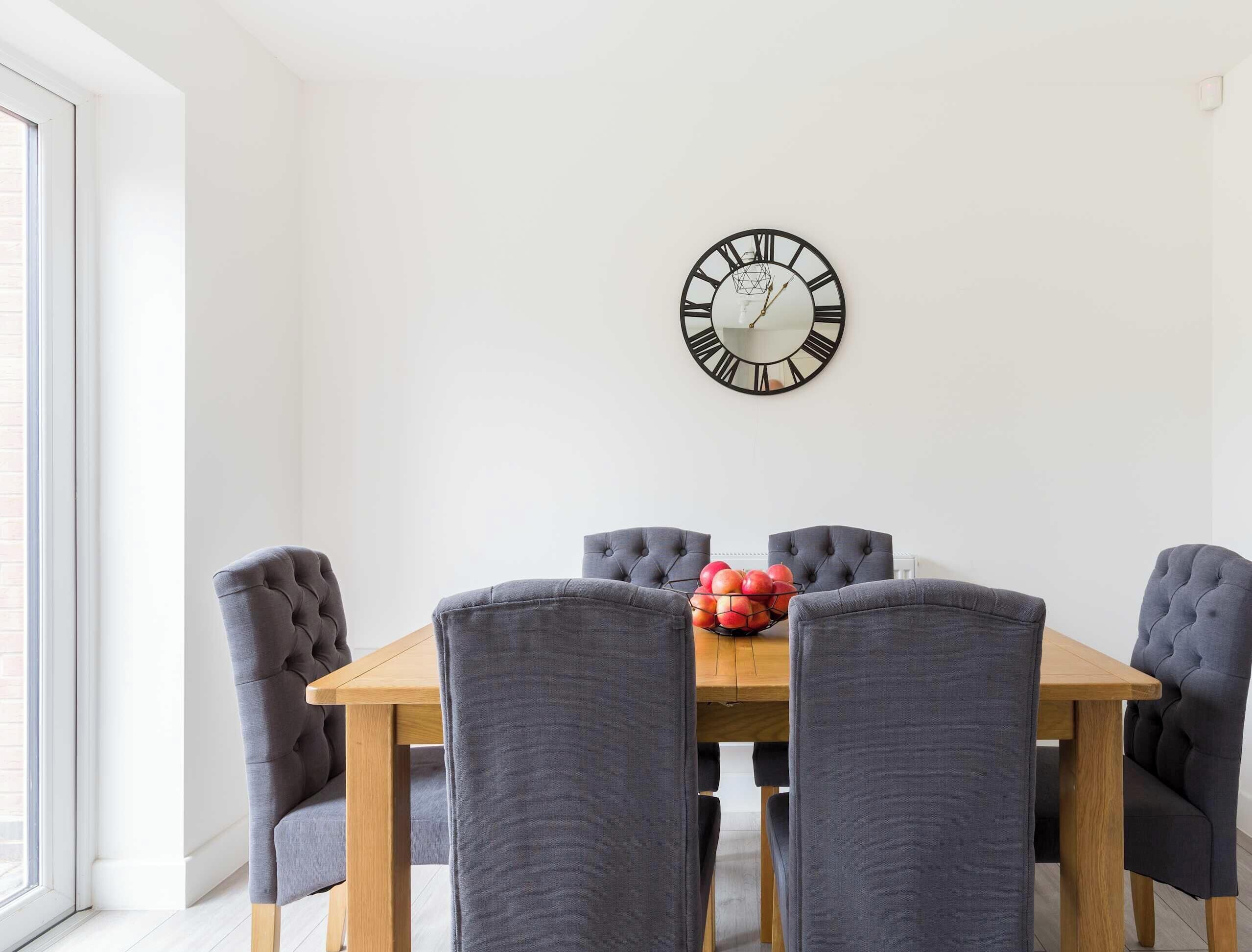A dining area with a wooden table, grey upholstered chairs, and a wall-mounted clock.