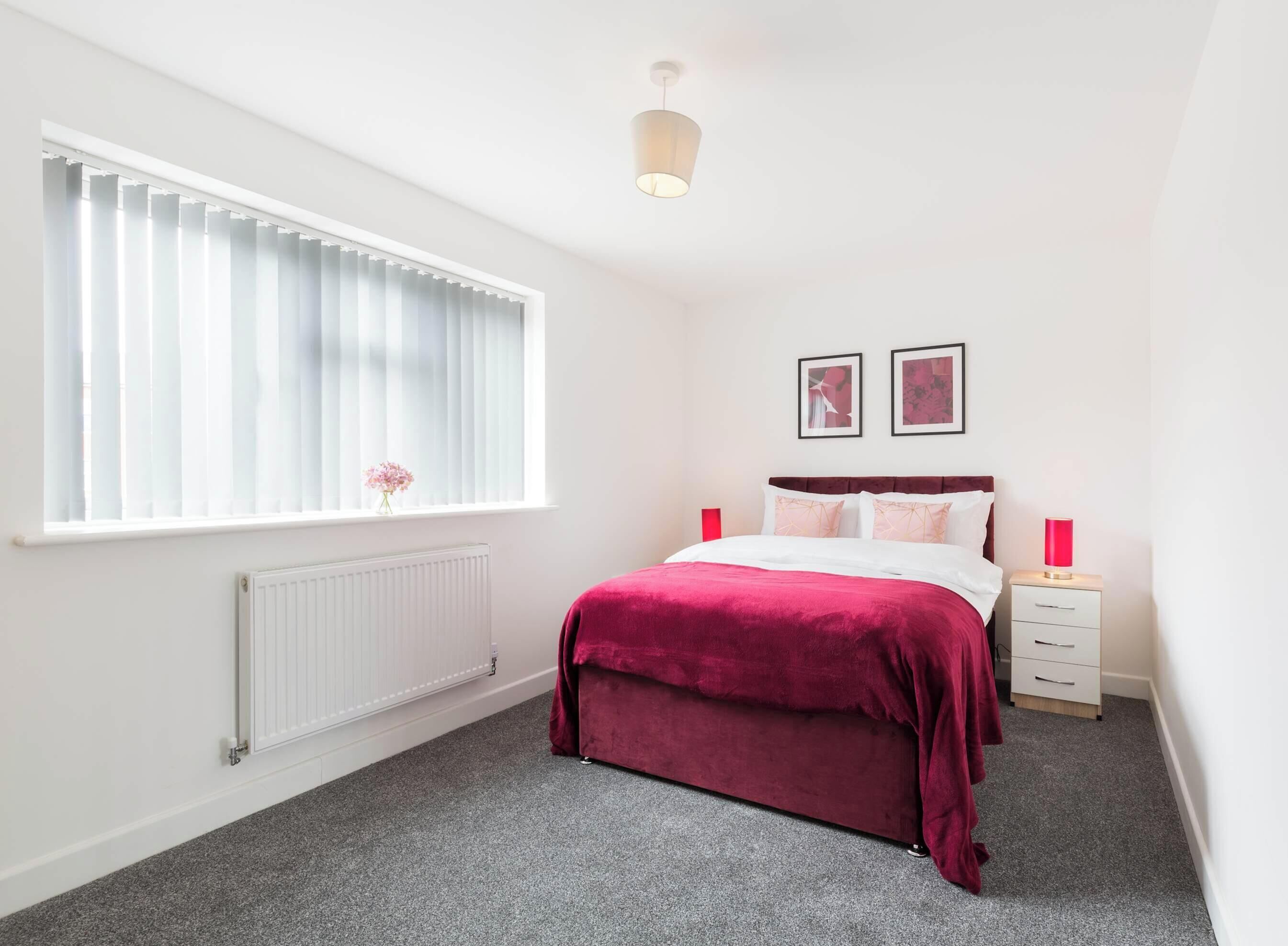 Een slaapkamer met een bed, een radiator, een raam met zonwering, een nachtkastje en twee ingelijste foto's aan de muur.
